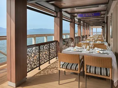 Das Restaurant Le Bistro an Bord der Norwegian Joy bietet Tische mit Meerblick. Die elegant gedeckten Tische stehen entlang einer Fensterfront mit Blick auf das offene Meer und schneebedeckte Berge. Ohne Personen.