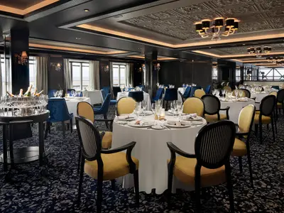 Elegantes Restaurant an Bord der Norwegian Joy. Zahlreiche festlich gedeckte Tische mit blauen und gelben Stühlen und ein Sektwagen. Große Fenster mit Meerblick.