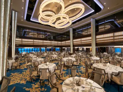 Das Restaurant an Bord der Norwegian Joy zeigt zahlreiche gedeckte Tische und Stühle. Große Fenster bieten Meerblick. Ring-Kronleuchter hängen von der Decke. Blauer Teppich mit goldenen Mustern.