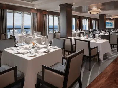 Das elegante Ocean Blue Spezialitätenrestaurant an Bord der Norwegian Joy. Große Fenster mit Meerblick. Zahlreiche schick gedeckte Tische mit weißen Tischdecken, Gläsern und Besteck im stilvollen Innenraum.