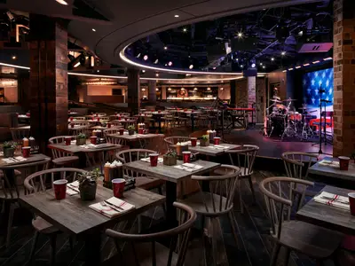 Ansicht des Restaurants Q an Bord der Norwegian Joy, mit zahlreichen Holztischen, Stühlen und einer Bühne mit Schlagzeug und American-Flag-Bildschirm. Gemütliche Abendstimmung.