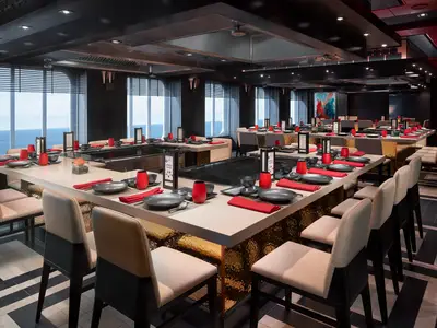 Blick in das stilvolle Teppanyaki-Restaurant an Bord der Norwegian Joy. Es gibt mehrere Kochstationen, Tische sind mit roten Servietten und dunklem Geschirr gedeckt. Große Fenster bieten einen weiten Blick auf das offene Meer.