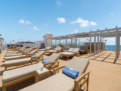 Blick auf das obere Sonnendeck der Norwegian Joy mit zahlreichen beigen Sonnenliegen und blauen Handtüchern. Im Hintergrund das blaue Meer und ein strahlender Himmel.