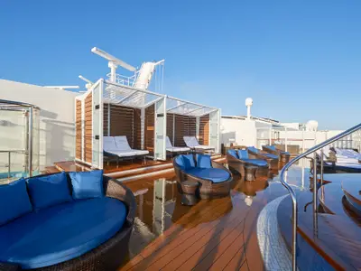 Ein sonniges Bild des The Haven Sonnendecks an Bord der Norwegian Joy. Es zeigt blaue Lounge-Möbel, weiße Sonnenliegen, eine Außendusche und ein glänzendes Holzdeck mit Reflexionen.