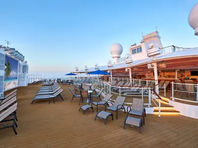 Das menschenleere Sonnendeck SpiceH2O der Norwegian Joy bei Abendstimmung. Liegestühle, Tische, blaue Schirme und eine Bar sind zu sehen, dazu eine Leinwand mit Strandmotiv.