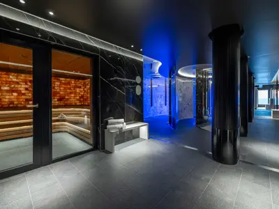 Moderner Spa-Bereich an Bord der Norwegian Joy mit einer hell beleuchteten Salzsauna, dunklen Marmorwaenden und blauen Erlebnisduschen. Mehrere Holzbaenke und gestapelte Handtuecher sind sichtbar.