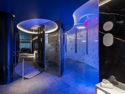 Ein futuristisches Spa an Bord der Norwegian Joy mit blauer und violetter Beleuchtung. Mehrere Erlebnisduschen sind in runden Nischen mit Marmorwänden sichtbar. Der Boden hat ein Mosaikmuster aus Kieselsteinen.