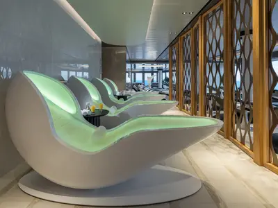 Moderne, beleuchtete Loungesessel in der Spa-Lounge der Norwegian Joy. Mit Meerblick durch große Fenster. Getränke stehen bereit. Elegantes Interieur mit Holz und Glas.
