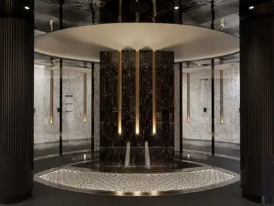 Ein luxuriöser Innenbereich der Thermal Suite an Bord der Norwegian Joy. Im Zentrum befindet sich ein "Pebble Walk" aus weissen Steinen, umgeben von einem dunklen Wasserbecken. Im Hintergrund sind dunkle Marmorwände mit vertikalen Leuchten und darüber eine runde Deckenverkleidung zu sehen. Grosse dunkle Säulen mit Rillen strukturieren den Raum.