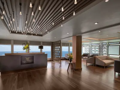 Empfangsbereich des Mandara Spa & Salon an Bord der Norwegian Joy. Moderne Rezeption, bequeme Loungebereiche mit Holzfußboden und Meerblick. Stylische Deckenleuchten und Produktregale sind sichtbar. Ohne Personen.