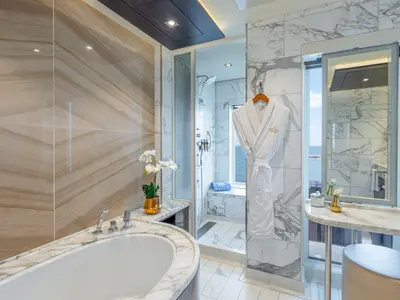 Badezimmer einer The Haven Suite an Bord der Norwegian Joy. Es zeigt eine Badewanne, eine begehbare Dusche, Marmorwände, eine goldene Vase mit Orchideen und einen Bademantel. Durch das Fenster ist das Meer zu sehen.