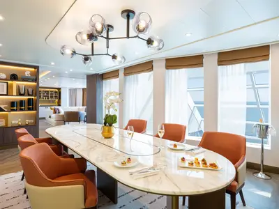 Ein luxuriöser Essbereich in der Haven Premier Owners Suite an Bord der Norwegian Joy. Ein langer Marmortisch ist gedeckt mit Häppchen und Sektgläsern. Große Fenster bieten einen weiten Blick auf das Meer. Im Hintergrund ist ein Wohnbereich und eine Bar sichtbar.