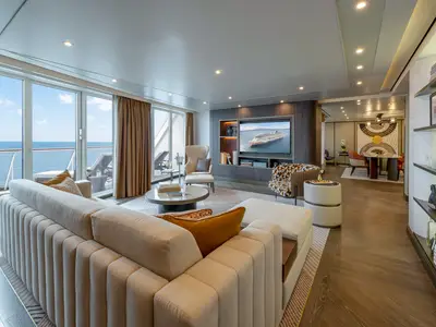 Grosses, luxuriöses Wohnzimmer einer Owners Suite an Bord der Norwegian Joy mit beigem Sofa, modernen Möbeln, einem grossen TV und Meerblick durch eine Panorama-Schiebetür.