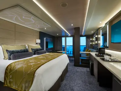 Eine elegante Suite an Bord der Norwegian Joy. Sie umfasst ein Doppelbett mit goldener Tagesdecke, einen Wohnbereich, einen Schreibtisch mit Kaffeemaschine und einen Balkon mit Meerblick.