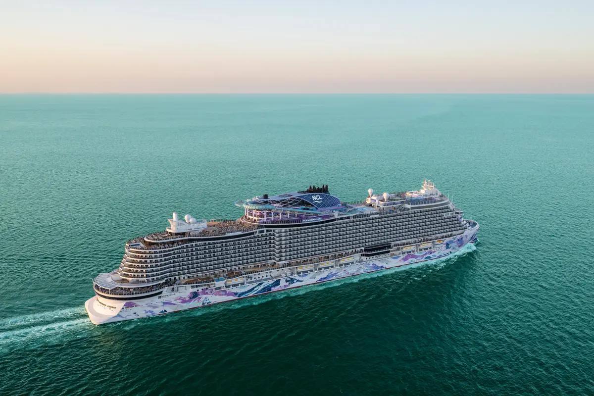 An aerial view of the Norwegian Luna cruise ship sailing on a turquoise sea. The ship features a modern, colorful hull design with purple and blue accents, cutting through the water under a pale sky.