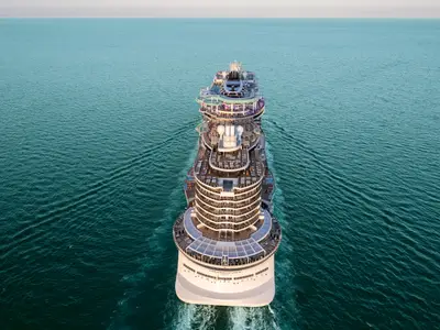 Luftaufnahme der Norwegian Luna auf dem offenen Meer. Das Megaschiff zeigt seine belebten oberen Decks mit zahlreichen Sonnenliegen, Pools und bunten Wasserrutschen. Der Ozean ist türkisblau und leicht wellig.