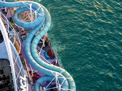 Vogelperspektive auf die gewundenen blauen Wasserrutschen auf dem Deck der Norwegian Luna. Das tiefe Meer daneben reflektiert die Sonne, was goldene Glitzerpunkte erzeugt.