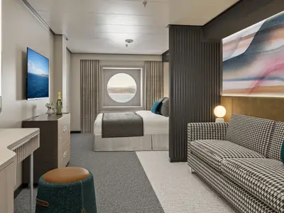Blick in eine geraeumige Aussenkabine der Norwegian Luna mit grossem Bullaugenfenster, Doppelbett, gemuetlichem Sofa und Schreibtisch. Der Raum ist modern und hell eingerichtet, mit Blick aufs Meer durch das Fenster.