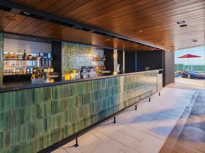 Die schicke Outdoor Bar Haven an Bord der Norwegian Luna mit einer Theke aus gruenen Hochglanzfliesen, gefuellt mit Spirituosen und Bierzapfanlagen. Im Hintergrund ist das Meer und ein roter Sonnenschirm zu sehen.