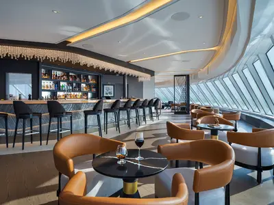 Elegante Bar-Lounge der Norwegian Luna. Lange Bar, Barhocker, Spirituosenregale, Kristall-Licht. Lederfauteuils, Tische mit Getränken. Große Schrägfenster mit Meerblick.