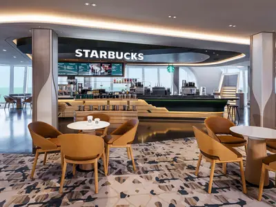 Starbucks Café an Bord der Norwegian Luna. Elegante Holzstühle und Tische auf gemustertem Teppich. Moderner Thekenbereich mit Maschinen und Beleuchtung. Panoramafenster bieten Meerblick. Zwei Starbucks-Becher auf einem Tisch.