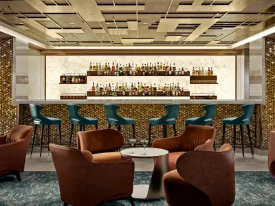 Eine moderne Bar an Bord der Norwegian Luna mit einer langen Theke, gefuellt mit unzähligen Flaschen, und stilvollen Sitzgelegenheiten in Teal und Braun. Der Raum strahlt eine elegante Abendstimmung aus.
