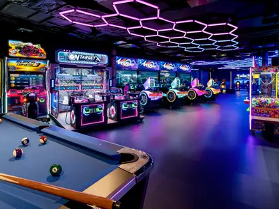 Die neonbeleuchtete Spielzone an Bord der Norwegian Luna zeigt Arcade-Fahrsimulatoren, Schiessspiele und einen Billardtisch. Lebhafte violette und blaue Lichter dominieren den Raum.