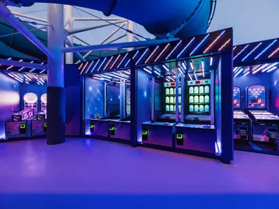 A vibrant arcade area on board the Norwegian Luna cruise ship, featuring multiple game stations with bright neon lights and a starry sky theme. The area is illuminated in blue and purple hues, with various interactive games ready for players, all without people present.