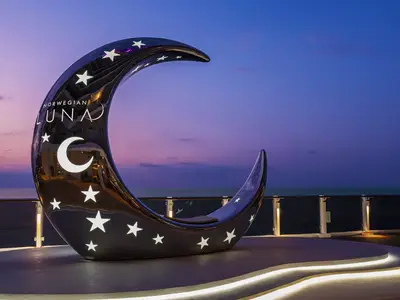 Ein großer, dunkler, glänzender Mond-Sessel mit leuchtenden Sternen und einem Mond-Symbol, der zum Sitzen einlädt, steht auf dem Deck der Norwegian Luna. Im Hintergrund ist das Meer und ein farbenfroher Abendhimmel zu sehen.