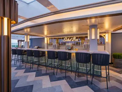 Die stilvolle Soleil Bar an Bord der Norwegian Luna. Eine lange Bar mit Hochstühlen, Neonlicht-Schriftzug und zahlreichen Getränkezapfhähnen. Im Hintergrund ist ein Teil des Außendecks sichtbar.