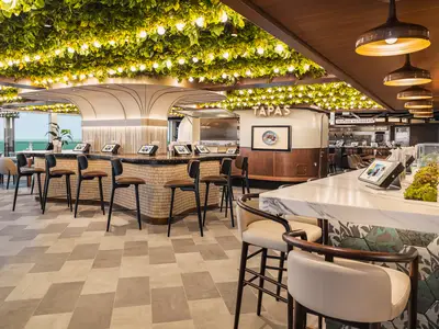 Moderne Indulge Food Hall an Bord der Norwegian Luna mit hellen Essensstationen und Meerblick. Grüne Deckenbepflanzung, digitale Menüs und Barhocker prägen den einladenden Raum.