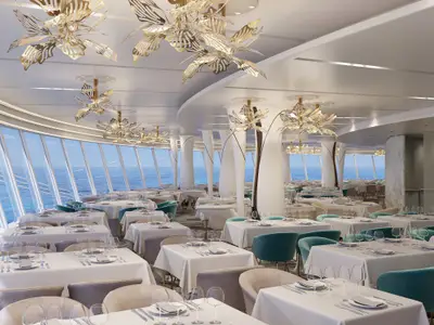 Hudson's Hauptrestaurant an Bord der Norwegian Luna. Der moderne Speisesaal hat Tische mit weißen Tischdecken, Stühlen und großen Fenstern mit Meerblick. Goldene Blütenleuchten zieren die Decke.