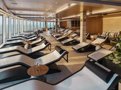 Innenansicht der Thermal Suite der Norwegian Luna. Moderne Liegen in Schwarz-Weiss stehen an Panoramafenstern mit Meerblick. Holzdetails und Pflanzen sorgen für entspanntes Ambiente.