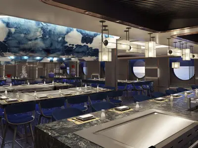 Das elegante Hasuki Teppanyaki Restaurant an Bord der Norwegian Luna. Blaue Stühle, dunkle Grilltische, moderne Hängelampen und eine kunstvolle blau-weiße Decke. Runde Fenster bieten Meerblick im stilvollen, menschenleeren Raum.