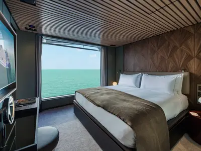 Ein modern eingerichtetes Schlafzimmer einer Suite an Bord der Norwegian Luna mit einem grossen Bett, weisser Bettwäsche und einem Panoramablick auf das Meer durch ein raumhohes Fenster. Links ist ein Fernseher zu sehen.