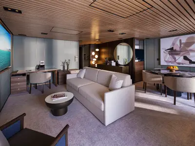 Ein heller, moderner Wohnbereich einer luxuriösen Suite an Bord der Norwegian Luna. Ein beiges Sofa, ein Esstisch mit braunem Marmor und ein Schreibtisch mit Stuhl sind zu sehen. Die Wände sind hellblau und dunkelbraun getäfelt, die Decke aus Holzlatten.