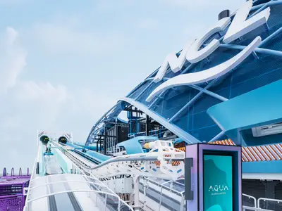 Außenansicht des Aqua Slide Coasters an Bord der Norwegian Luna. Die hellblaue Rutsche windet sich über das Deck, im Hintergrund das NCL-Logo und ein Display mit 'AQUA SLIDECOASTER'.
