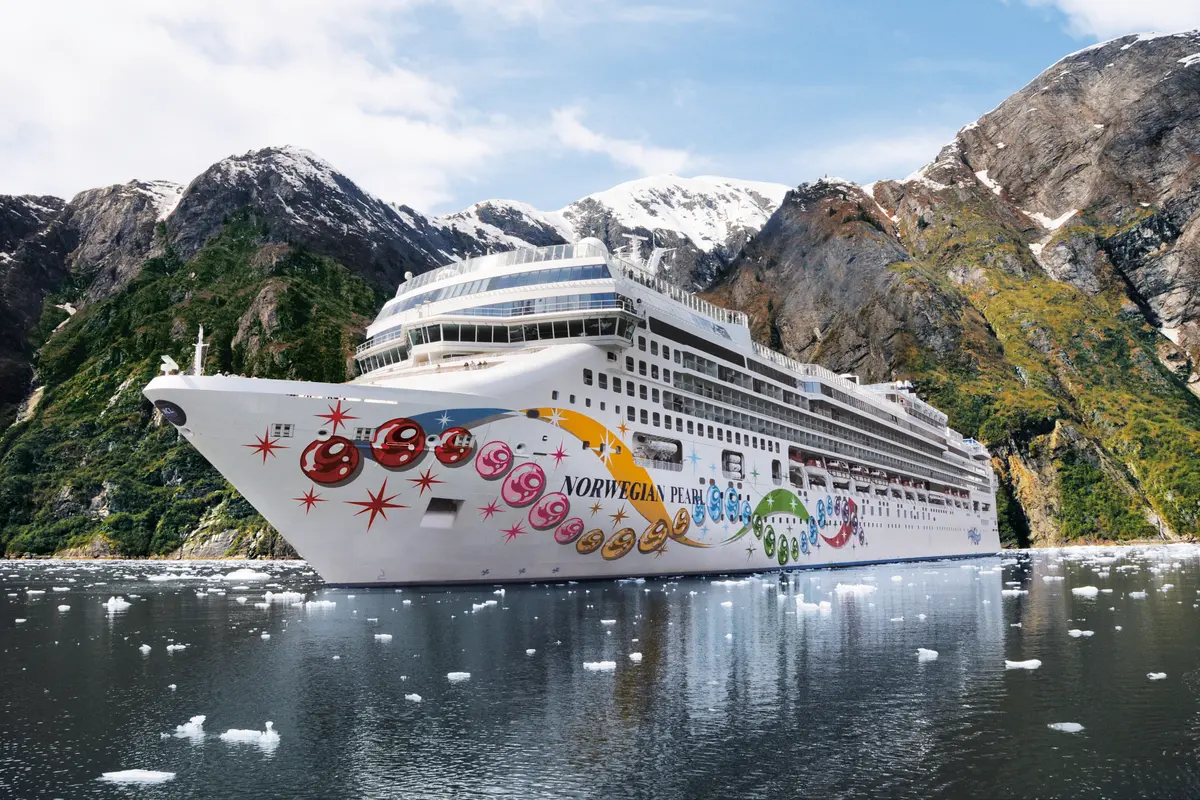 Die Norwegian Pearl, ein Kreuzfahrtschiff mit bunter Rumpfbemalung, faehrt durch einen eisigen Fjord. Schneebedeckte, bewaldete Berge umgeben das Schiff, und Eisbrocken schwimmen auf dem dunklen Wasser.