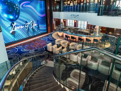 Blick von oben auf das Atrium an Bord der Norwegian Pearl. Zu sehen ist eine geschwungene, beleuchtete Bar mit Hockern, mehrere Sitzgruppen aus weissen Ledersesseln und Beistelltischen sowie eine grosse digitale Anzeigetafel mit abstrakter blauer Welle und Schriftzug 'Live it Up'. Eine geschwungene Treppe mit Glasgeländer führt nach unten.