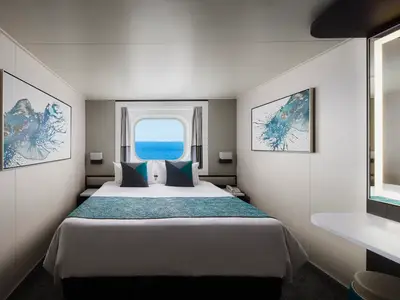 Eine helle Aussenkabine an Bord der Norwegian Pearl. Ein grosses Bullauge bietet Meerblick. Das Bett ist mit einer türkisfarbenen Tagesdecke und Kissen dekoriert. Zwei abstrakte Kunstwerke hängen an den Wänden.