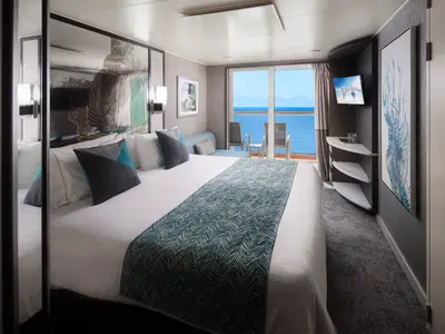 Eine helle Balkonkabine an Bord der Norwegian Pearl mit einem frisch gemachten Doppelbett, einem teal-farbenen Läufer und einem Sofa am Fenster. Im Hintergrund ist das blaue Meer zu sehen.