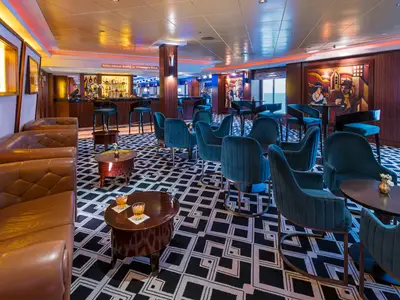 Blick in die Maltings Bar an Bord der Norwegian Pearl. Die Szene zeigt dunkle Holzvertäfelung, blaue Samtsessel, braune Ledersofas und eine beleuchtete Bar mit Kunstwerken.