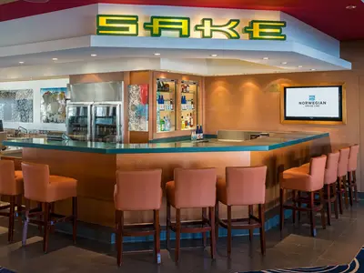 Blick in die helle Bar-Lounge Sake Bar an Bord der Norwegian Pearl mit leeren Barhockern, einer Theke und Flaschen hinter Glas. Ein Bildschirm zeigt das Logo von Norwegian Cruise Line.