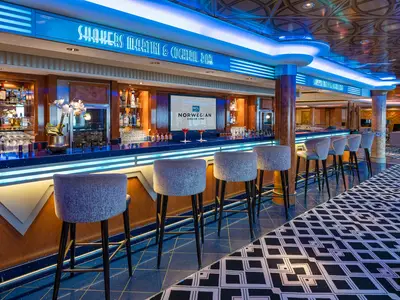 Die Shakers Martini & Cocktail Bar an Bord der Norwegian Pearl. Eine moderne, beleuchtete Bar mit Spirituosen, Gläsern und Cocktails. Mehrere graue Barhocker stehen davor. Ein Bildschirm zeigt das NCL-Logo.