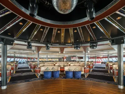Die Spinnaker Lounge an Bord der Norwegian Pearl. Geräumige Bar-Lounge mit gestuften Sitzgelegenheiten. Moderne Sofas, blaue Hocker und Bühne mit Beleuchtung. Bar und Fenster mit Meerblick im Hintergrund.