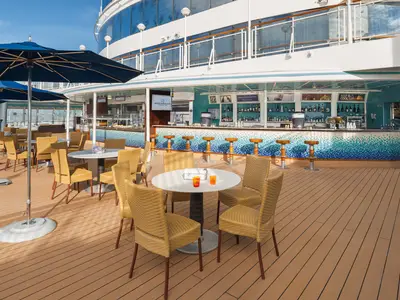 Ein lebhafter Außenbereich an Bord der Norwegian Pearl. Die Topsiders Bar mit blauen Mosaikfliesen und Barhockern. Davor Tische, Korbstühle und Sonnenschirme auf dem Teakdeck.