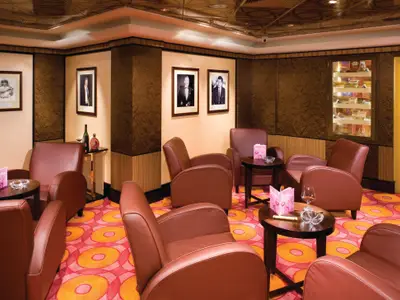 Ein stilvoller, gemütlicher Cigar Club an Bord der Norwegian Pearl mit bequemen braunen Ledersesseln, kleinen Tischen mit Getränken und einem Zigarren-Humidor an der Wand. Der Teppich hat ein leuchtendes, rotes und orangefarbenes Muster.
