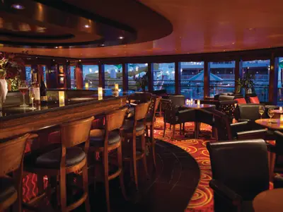 Die Star Bar der Norwegian Pearl: dunkle Holztheke, Barhocker und Sitzgruppen. Blick auf beleuchtetes Pooldeck mit Wasserrutschen bei Abendstimmung.