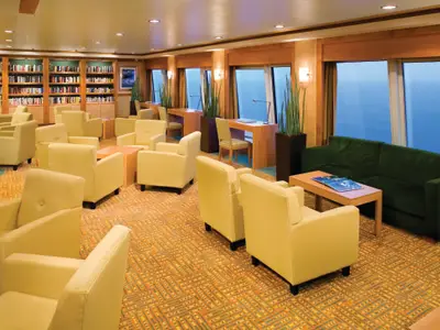 Die helle Bibliothek der Norwegian Pearl bietet zahlreiche gelbe Sessel, dunkelgrüne Sofas und Schreibtische. Bücherregale füllen eine Wand, während große Fenster einen weiten Meerblick offenbaren.