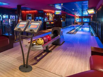 Eine modern beleuchtete Bowlingbahn in der Bliss Ultra Lounge an Bord der Norwegian Pearl. Drei Bowlingkugeln, darunter eine mit Smiley-Gesicht, liegen bereit. Blaue und rote Lichteffekte dominieren den Raum.